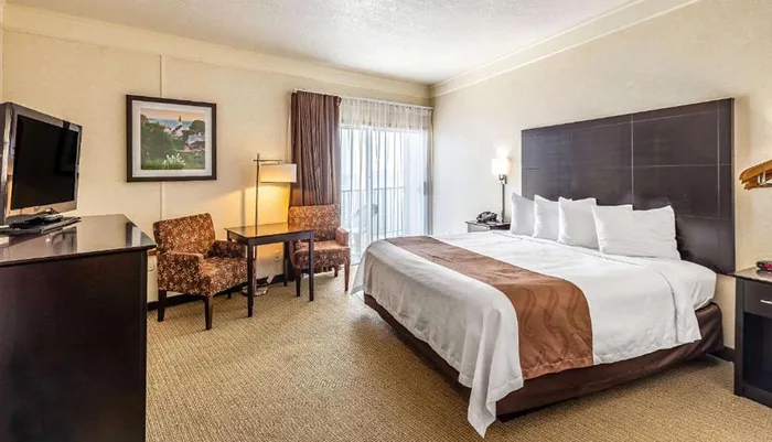 The image shows a tidy hotel room with a large bed two upholstered chairs a work desk a flat-screen TV and a framed picture on the wall