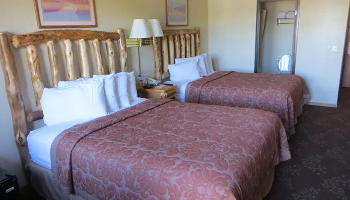 The image shows a hotel room with two beds featuring rustic wooden headboards patterned bedspreads a side table with a lamp and a wall mirror reflecting part of the room