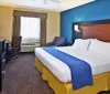The image displays a neatly arranged hotel room featuring a king-sized bed with white linens a blue and yellow color scheme and typical furnishings like a desk chair and a flat-screen TV