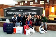A group of people is posing with shopping bags and luggage in front of the Woodbury Common Premium Outlet sign, seemingly happy after a shopping spree.