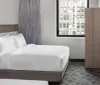 The image depicts a neatly made bed with white linens in a modern hotel room with a view of the adjacent building through the window
