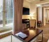 The image shows a neatly arranged hotel room with a bed desk and television featuring a view into an adjoining room with seating areas