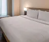 The image shows a neatly made bed with white linens in a tidy modern hotel room with natural light coming through sheer curtains