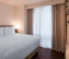 The image shows a neatly made bed with white linens in a tidy modern hotel room with natural light coming through sheer curtains