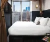 A neatly made bed with white linens is set against a dark blue headboard with a large window showcasing an urban skyline
