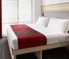 The image shows a neatly made bed with white linens and a red bed runner in a modern hotel room with a work desk and closed blinds on the window