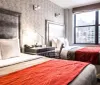 The image shows a modernly decorated hotel room with a large bed featuring a decorative red throw a smaller bed elegant furnishings and a view of urban buildings through the window