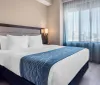 The image shows a neatly arranged hotel room with a large bed crisp white bedding a blue decorative throw flanked by bedside tables and lamps with a large window providing a view of the outside urban area