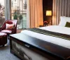 The image shows a modern and elegant hotel room with a comfortable bed stylish furniture and a balcony with a city view