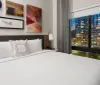 The image shows a neatly made bed in a modern hotel room with artistic wall decor and a large window offering a view of a cityscape at night