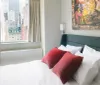 A modern bedroom with a simple design features a neatly made bed with white sheets and red pillows an abstract painting on the wall and a window view of urban skyscrapers