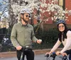 Two people wearing helmets and sunglasses are smiling while riding bicycles by a flowering tree on a sunny day