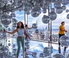 Visitors explore a reflective art installation filled with silvery spheres against the backdrop of a high-rise cityscape seen through large windows