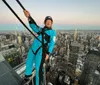 Two people in blue jumpsuits are suspended by safety harnesses high above a cityscape with one of them raising their arms in excitement