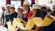 A group of individuals, wearing white chef hats, appear engaged and happy as they participate in a cooking class or culinary workshop.