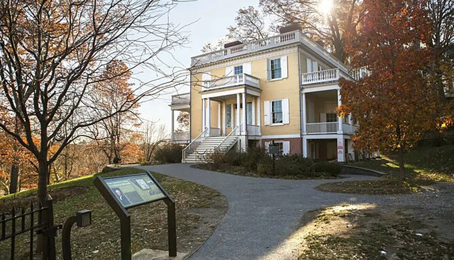 The image shows a stately yellow house with white columns and balconies, situated on a leaf-covered lawn with bare trees and an information plaque in the foreground, in the gentle light of either sunrise or sunset.