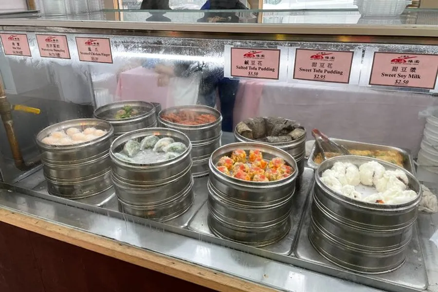 The image displays a variety of Chinese dim sum dishes in traditional steaming baskets, set on a counter for sale, with price tags for each dish visible in the background.