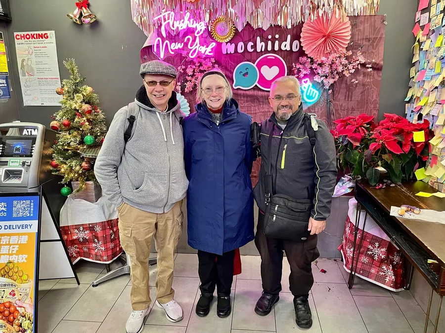 Three people are smiling for a photo inside a festive-looking shop decorated with Christmas and floral elements, including a sign that reads Flushing New York mochido.