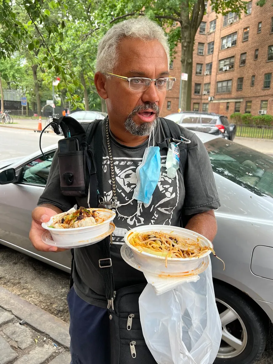A man with glasses and a face mask dangling from his ear looks surprised while holding two plates of food outdoors.