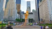 The image shows a vibrant city scene featuring the gilded statue of William Tecumseh Sherman at Grand Army Plaza, with a bustling crowd and urban high-rises in the backdrop.