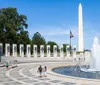 The image shows the World War II Memorial with its fountain in the foreground and the Washington Monument in the background on a sunny day with visitors walking around and enjoying the scenery