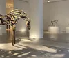 The image shows a gallery space featuring an art installation with sculptures of horses made from branches and wood positioned as if in motion