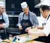Two men wearing chef hats and aprons watch attentively as a professional chef demonstrates a culinary technique