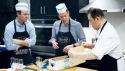Two men wearing chef hats and aprons watch attentively as a professional chef demonstrates a culinary technique.