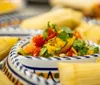 A colorful dish with fresh chopped vegetables is in focus set against a background with out-of-focus tamales wrapped in corn husks