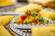 A colorful dish with fresh chopped vegetables is in focus, set against a background with out-of-focus tamales wrapped in corn husks.