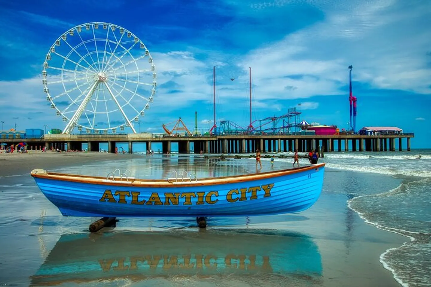 A lifeboat with the words Atlantic City painted on its hull is positioned on a sandy beach against the backdrop of a pier with amusement park rides, including a Ferris wheel, under a partly cloudy sky.