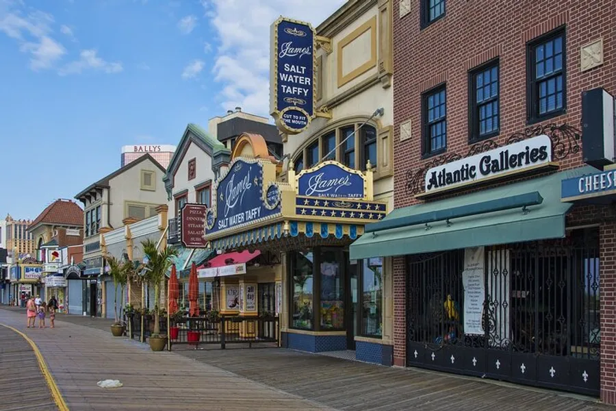 The image shows a sunny boardwalk lined with colorful storefronts, including one advertising salt water taffy, with a few people strolling by.