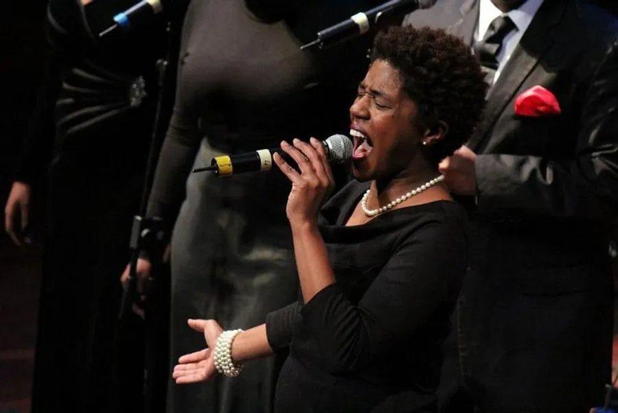 A woman passionately sings into a microphone with a choir in the background.