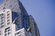 The image depicts the upper section of an Art Deco skyscraper with distinctive terraced crown and decorative metalwork against a clear blue sky.