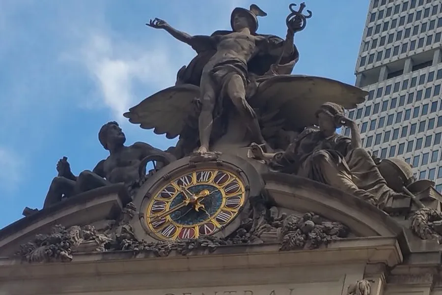 The image shows an ornate clock and sculptures atop the entrance of Grand Central Terminal in New York City against a backdrop of a clear sky and a modern building.