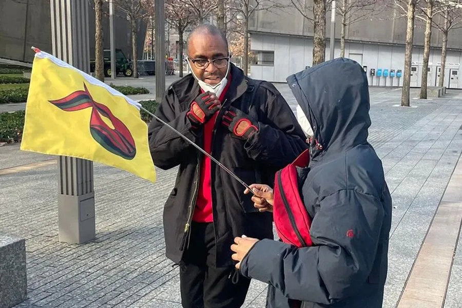 Two individuals, one holding a flag with a red and yellow emblem, are engaged in a conversation outdoors.