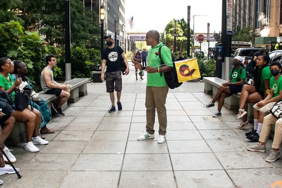 A person in a green shirt appears to be speaking to a group of attentive listeners outdoors near a city street.