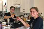 Two people are cheerfully toasting with cocktails in a home kitchen setting.