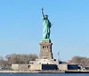The image shows the Statue of Liberty under a clear blue sky seen from a distance with water in the foreground