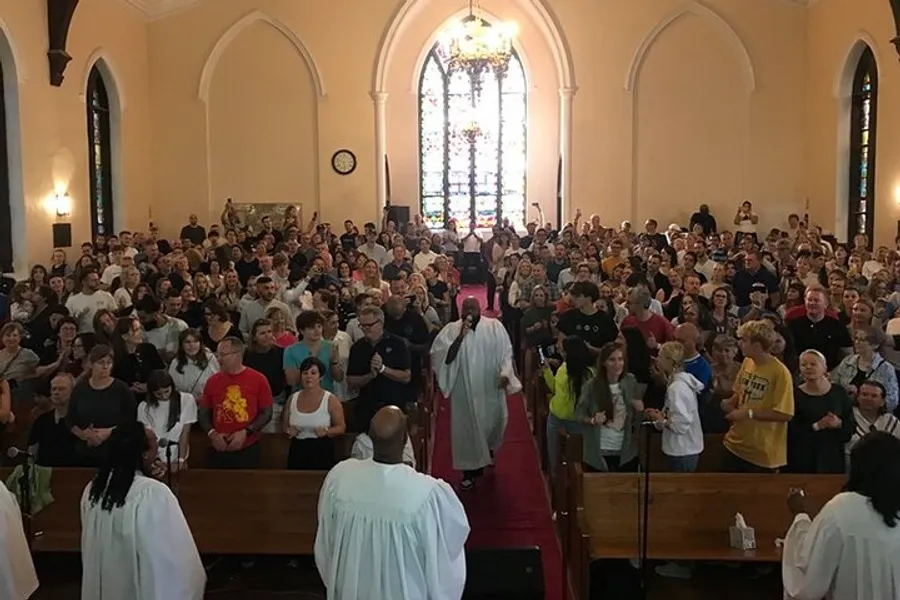 The image shows a congregation of people inside a church, standing as they face the aisle with a clergy member walking down the center.