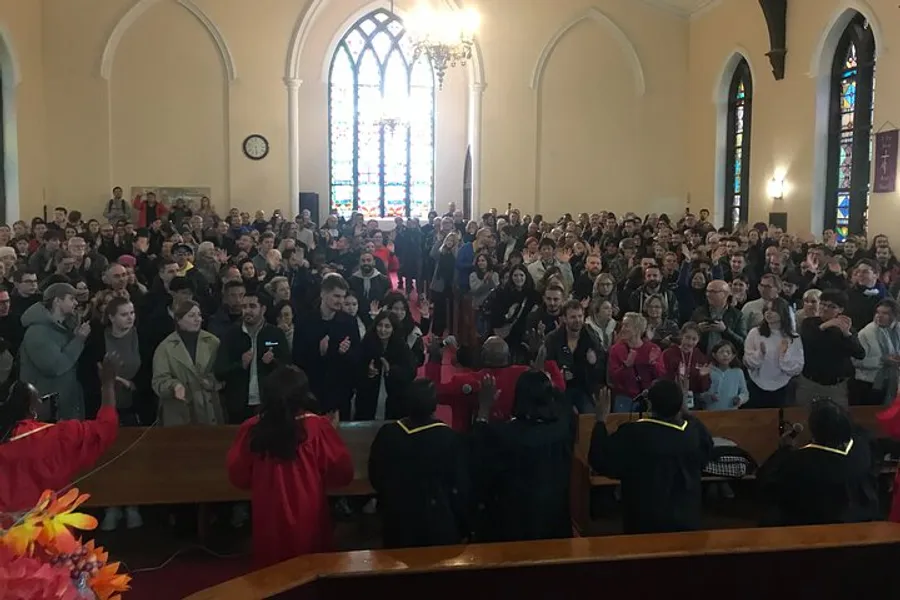 A diverse group of people is gathered inside a church, clapping and appearing to be engaged in a celebratory or significant event.