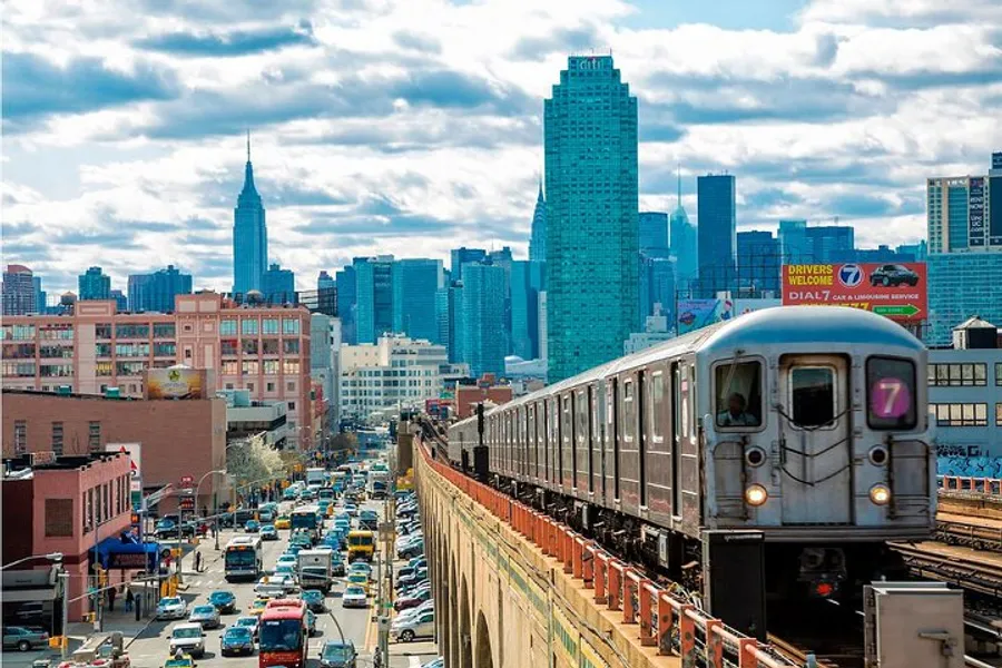 A train travels on elevated tracks above a bustling street with the New York City skyline in the background.