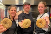 Three smiling individuals in a kitchen setting proudly hold up large, seed-covered bagels towards the camera.