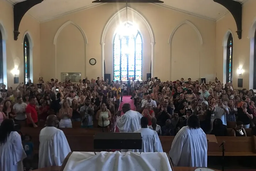 The image shows a congregation of people inside a church applauding, with some individuals standing at the front of the church dressed in white robes.