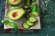 A halved avocado, sliced cucumber, jalapeño slices, and basil leaves are scattered on parchment paper with salt crystals, creating a fresh vegetable still life on a rustic wooden table.