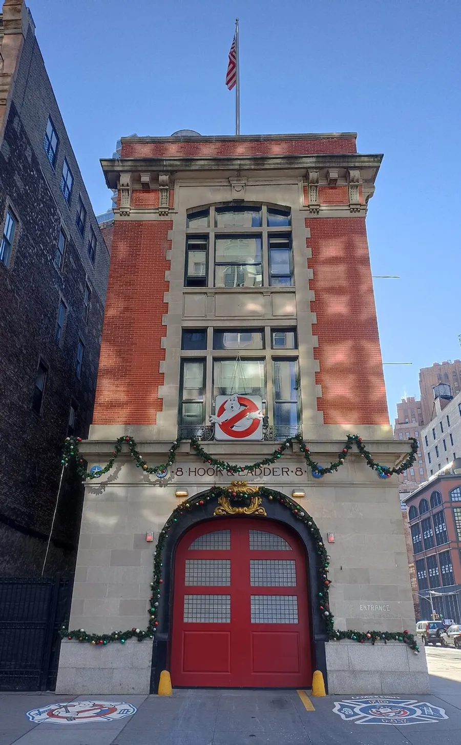 The image shows the exterior of a firehouse, adorned with Ghostbusters logo and holiday decorations, under a clear blue sky.