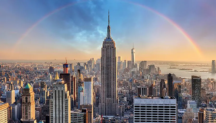 A panoramic view of New York City's skyline with a prominent view of the Empire State Building, under a wide rainbow arching across the scene.