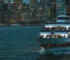 A cruise ship filled with passengers sails in front of a brightly lit city skyline during twilight hours