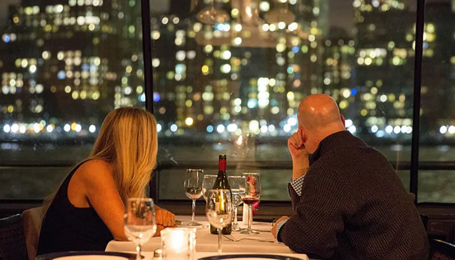 A man and woman sit at a dinner table by a window overlooking a city skyline at night.