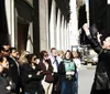 A group of people listens attentively as a man in a black jacket animatedly speaks or performs possibly on a city street or during a walking tour